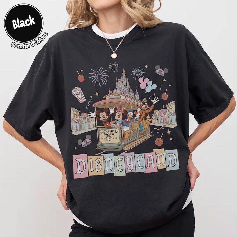 K&ouml;nnte beinhalten: Schwarzes T-Shirt mit einem Disneyland Main Street Trolley-Design. Die Grafik zeigt Micky Maus, Minnie Maus, Donald Duck und Goofy auf der Stra&szlig;enbahn, mit Feuerwerk und dem Schloss im Hintergrund. Das Wort "Disneyland" steht unten.