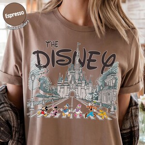 May include: An espresso-colored t-shirt featuring a graphic print of a castle and the text "THE DISNEY". Below the castle, Mickey Mouse, Minnie Mouse, Donald Duck, Daisy Duck, Goofy, and Pluto are walking across a crosswalk in a line.