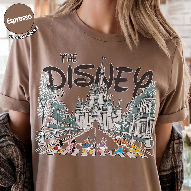 May include: An espresso-colored t-shirt featuring a graphic print of a castle and the text "THE DISNEY". Below the castle, Mickey Mouse, Minnie Mouse, Donald Duck, Daisy Duck, Goofy, and Pluto are walking across a crosswalk in a line.