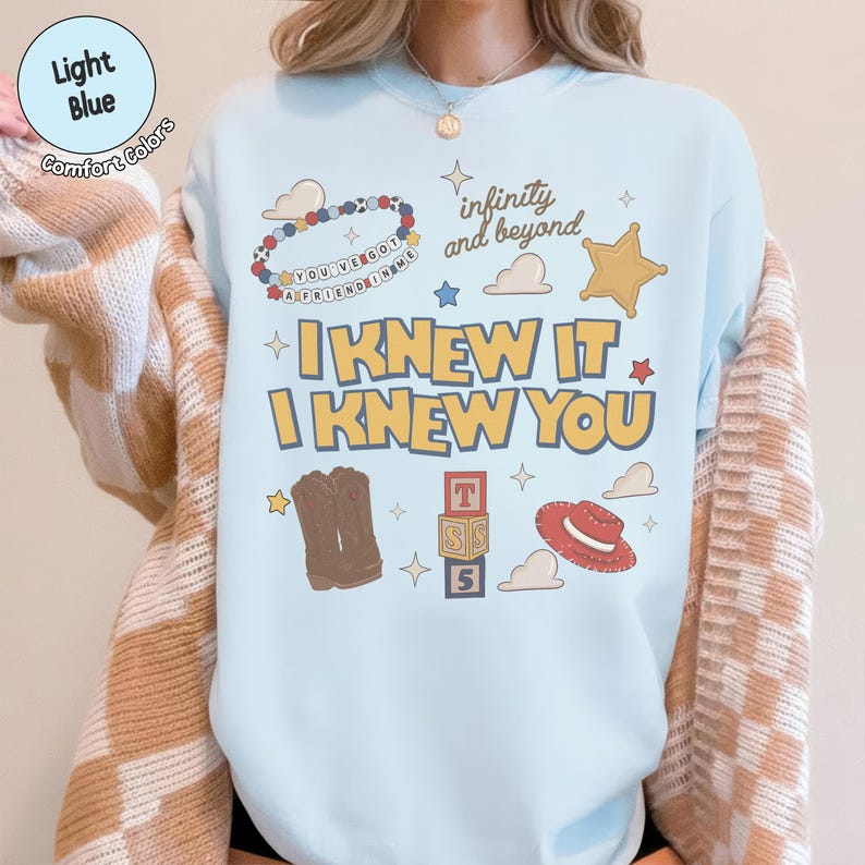Puede incluir: Una sudadera de cuello redondo azul claro con un estampado gr&aacute;fico que incluye el texto "I KNEW IT I KNEW YOU", "infinity and beyond" y "YOU'VE GOT A FRIEND IN ME". El dise&ntilde;o incluye ilustraciones de una pulsera de cuentas, botas vaqueras, bloques y un sombrero.