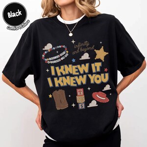 Puede incluir: Una camiseta negra con un estampado gr&aacute;fico que incluye el texto "I KNEW IT I KNEW YOU", "infinity and beyond" y "YOU'VE GOT A FRIEND IN ME". El dise&ntilde;o presenta ilustraciones de una pulsera de cuentas, una estrella dorada, botas marrones, bloques y un sombrero rojo.
