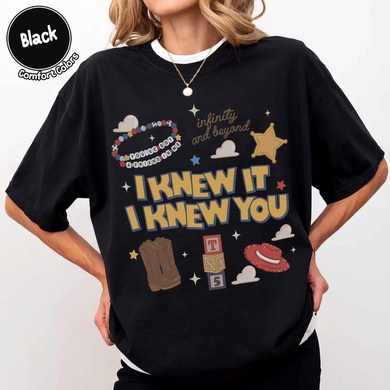 Puede incluir: Una camiseta negra con un estampado gr&aacute;fico que incluye el texto "I KNEW IT I KNEW YOU", "infinity and beyond" y "YOU'VE GOT A FRIEND IN ME". El dise&ntilde;o presenta ilustraciones de una pulsera de cuentas, una estrella dorada, botas marrones, bloques y un sombrero rojo.