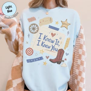 May include: A light blue crewneck sweatshirt featuring a central graphic of a lined notepad page with the text I Knew It, I Knew You. The design includes a brown cowboy boot, a wagon wheel, a sheriff star, hearts, stars, and tape strips.