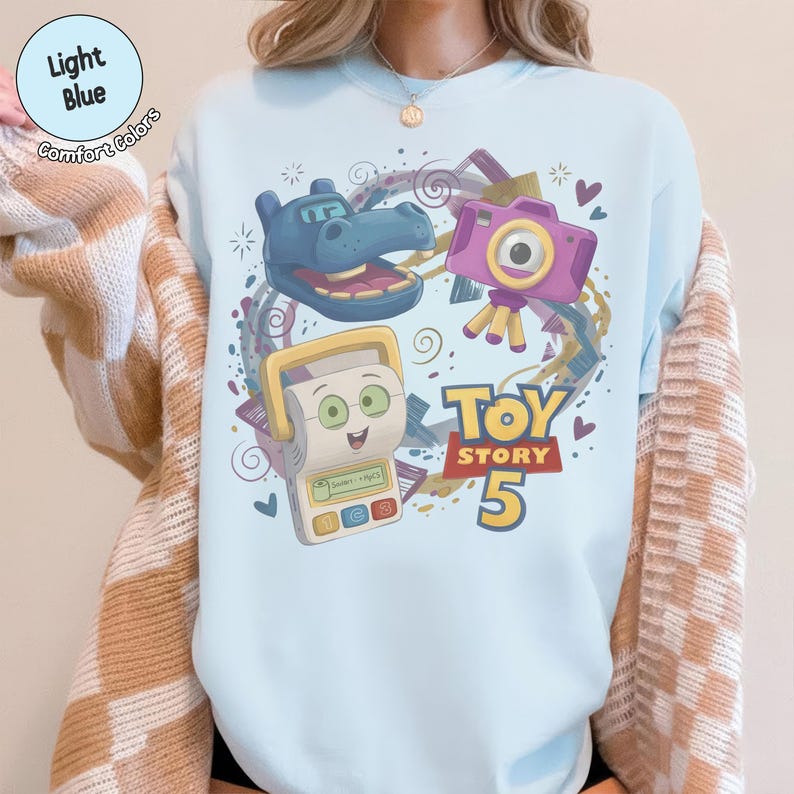 May include: A light blue crewneck sweatshirt featuring a graphic print of a blue hippo, a purple camera, and a white toy device with a screen and buttons. The text Toy Story 5 is printed in yellow and red. The shirt is layered under a tan and white cardigan.