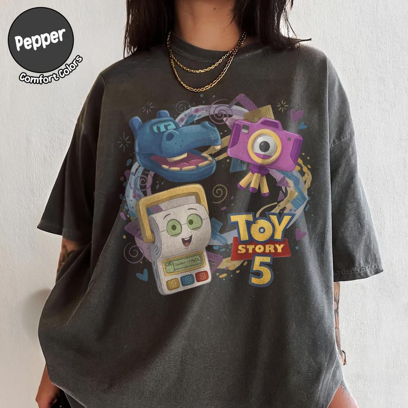 May include: A charcoal gray t-shirt featuring a graphic print of characters from Toy Story 5, including a blue hippo, a purple camera, and a white electronic toy. The text Toy Story 5 is printed in yellow and red. A Pepper Comfort Colors logo is visible.