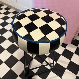 May include: Round stool featuring a black and white chequered seat, edged with blue piping. The seat is supported by a black metal frame. The stool is positioned on a black and white chequered floor.