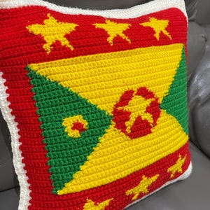 May include: Crocheted pillow featuring the flag of Grenada. The pillow has a red background with yellow stars, a yellow and green central design, and a white border. The design is a handmade, decorative item.
