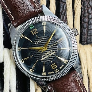 May include: A vintage Oris watch with a black face, gold hands, and a brown leather strap. The watch face features gold hour markers and the text "17 JEWELS SHOCK PROOF". The watch has a silver-colored bezel and a brown leather strap.