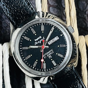 Op de afbeelding: Een vintage HMT Rajat automatisch polshorloge met een zwarte wijzerplaat en een zwarte leren band. De wijzerplaat toont de dag en datum, met een rode secondewijzer. Het horloge heeft een zilverkleurige kast.