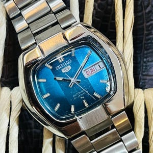 Peut inclure: Une montre automatique Seiko 5 vintage avec un boîtier carré argenté et un cadran bleu. Le cadran affiche le jour et la date, avec le jour indiquant "DIM 1". La montre a un bracelet en métal argenté.