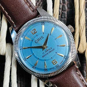 Może przedstawiać: Zegarek vintage z jasnoniebieską tarczą, złotymi znacznikami godzin i marką "CRIS". Zegarek ma srebrną ramkę i brązowy skórzany pasek. Tarcza zegarka zawiera również napis "17 JEWELS SHOCK PROOF".