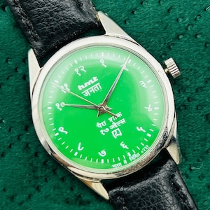 May include: A vintage wristwatch with a bright green face and silver-tone case. The watch features black hands, Arabic numerals, and text in a foreign language. It has a black leather band and a crown on the side.