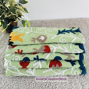 May include: A stack of four light green embroidered textiles. Each textile features a floral design with white, red, yellow, brown, and blue accents. The text "AnokhiCreationStore" is visible at the bottom.