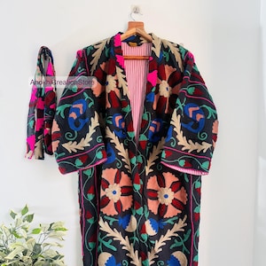 Puede incluir: Un abrigo largo negro bordado con una bufanda a juego. El abrigo presenta un diseño floral en tonos rojos, rosas, azules y dorados. El forro interior es de rayas rosas y blancas. La bufanda tiene un patrón floral similar. El abrigo está colgado en una percha de madera.