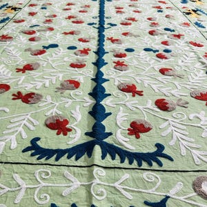 May include: A light green textile with intricate embroidery. The design features white, blue, red, and gray floral and leaf patterns. The text "AnokhiCreationStore" is visible in the lower right corner. The textile is likely a decorative item.