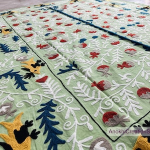 May include: A light green textile with detailed embroidery. The design includes white, red, blue, and yellow floral and leaf patterns. The textile has a black border and the text "AnokhiCreationStore".