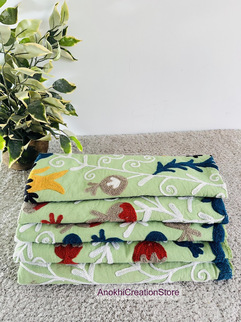 May include: A stack of four light green embroidered textiles. Each textile features a floral design with white, red, yellow, brown, and blue accents. The text "AnokhiCreationStore" is visible at the bottom.