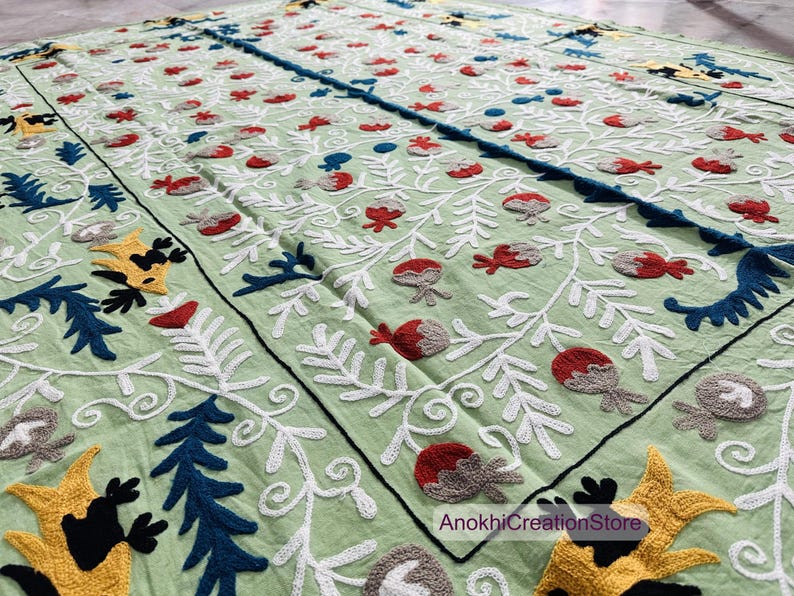 May include: A light green textile with detailed embroidery. The design includes white, red, blue, and yellow floral and leaf patterns. The textile has a black border and the text "AnokhiCreationStore".