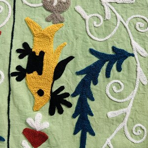 May include: Close-up of embroidered fabric with a light green background. The design features a yellow fish-like figure with black accents, blue leaf-like shapes, and white floral patterns. The text "AnokhiCreationStore" is visible.