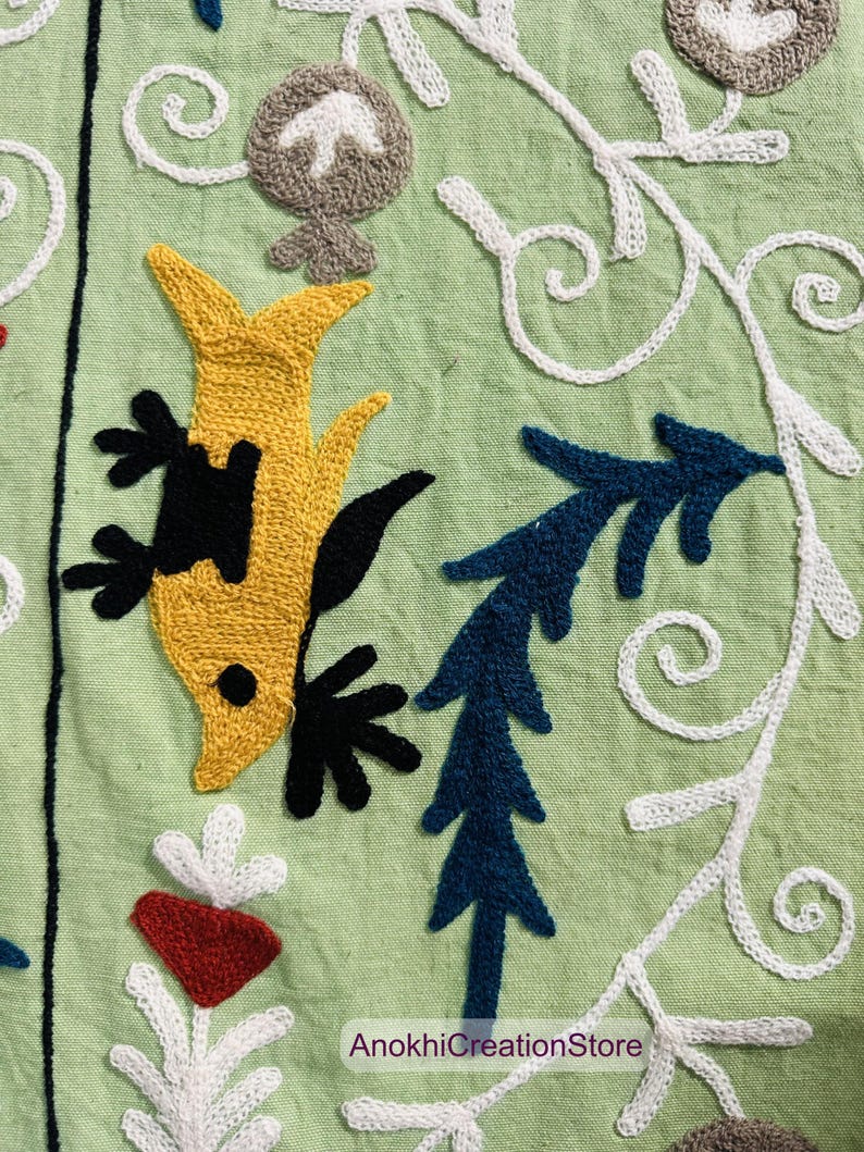 May include: Close-up of embroidered fabric with a light green background. The design features a yellow fish-like figure with black accents, blue leaf-like shapes, and white floral patterns. The text "AnokhiCreationStore" is visible.