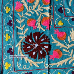 May include: Close-up of a teal textile with intricate embroidery. The design features floral and botanical motifs in shades of pink, orange, yellow, and brown, with cream-colored accents. The text "AnokhiCreationStore" is visible.