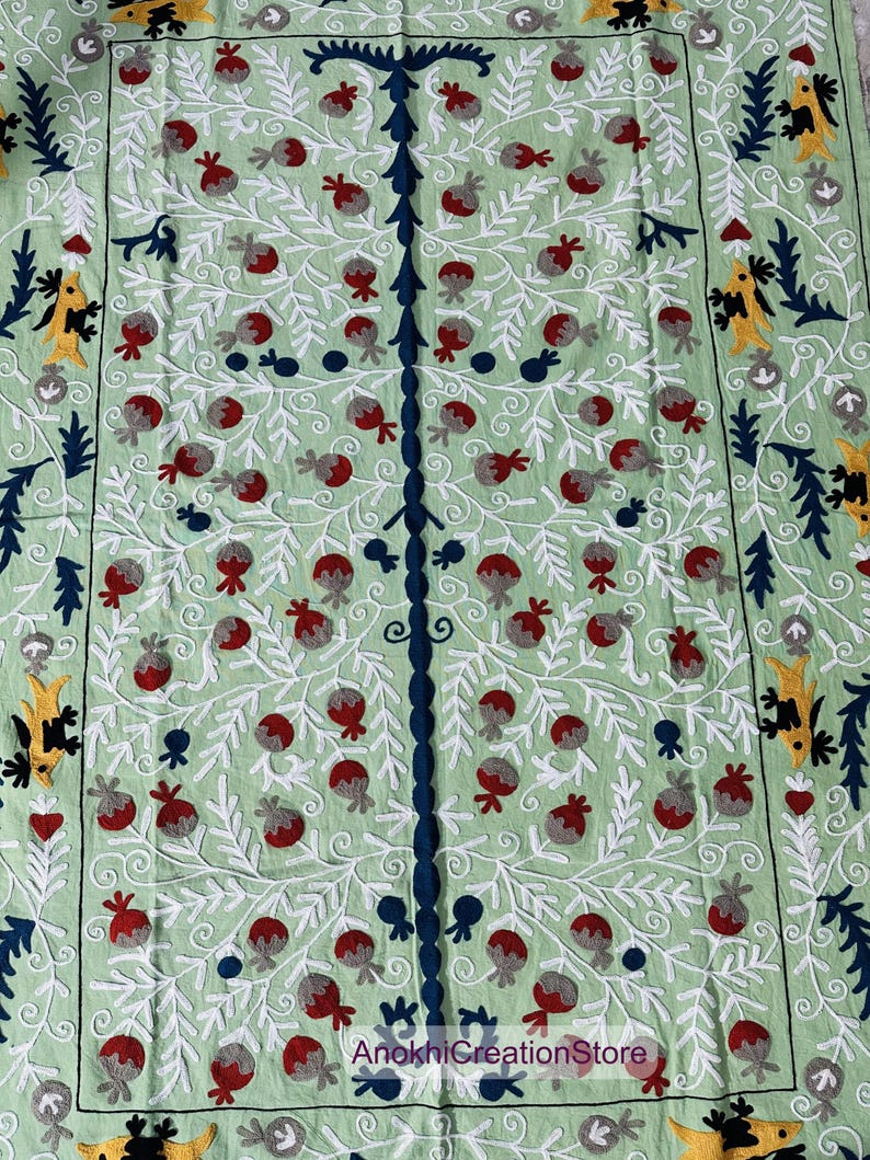 May include: A light green textile with an embroidered tree design. The tree has a dark blue trunk and branches, with red and gray fruit. White, yellow, and dark blue decorative elements are in the corners and edges. The text "AnokhiCreationStore" is at the bottom.