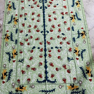 May include: A light green rectangular textile with an embroidered tree design. The tree has a dark blue trunk and branches, with red and gray fruit. The border features yellow and blue stylized bird and leaf motifs, with white floral accents.
