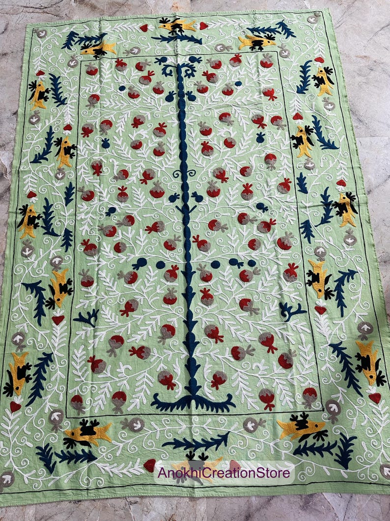 May include: A light green rectangular textile with an embroidered tree design. The tree has a dark blue trunk and branches, with red and gray fruit. The border features yellow and blue stylized bird and leaf motifs, with white floral accents.