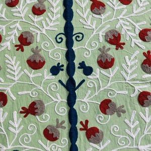 May include: Close-up of a light green textile with intricate embroidery. The design features white, stylized branches with red and gray fruit-like shapes. A central, dark blue embroidered column adds a contrasting vertical element. The text "AnokhiCreationStore" is visible.
