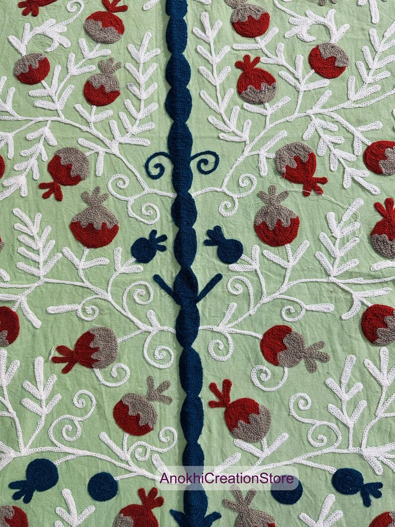 May include: Close-up of a light green textile with intricate embroidery. The design features white, stylized branches with red and gray fruit-like shapes. A central, dark blue embroidered column adds a contrasting vertical element. The text "AnokhiCreationStore" is visible.