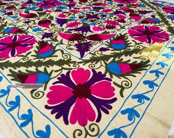 May include: Cream-colored textile with vibrant floral embroidery. The design features large pink and purple flowers, blue accents, and green foliage. A blue border with floral motifs frames the central pattern. The textile is likely a decorative item.