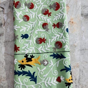 May include: A folded, light green textile with intricate embroidery. The design features white vines, red and gray floral elements, and yellow and black fish-like figures. The border is dark blue, and the text "AnokhiCreationStore" is visible.