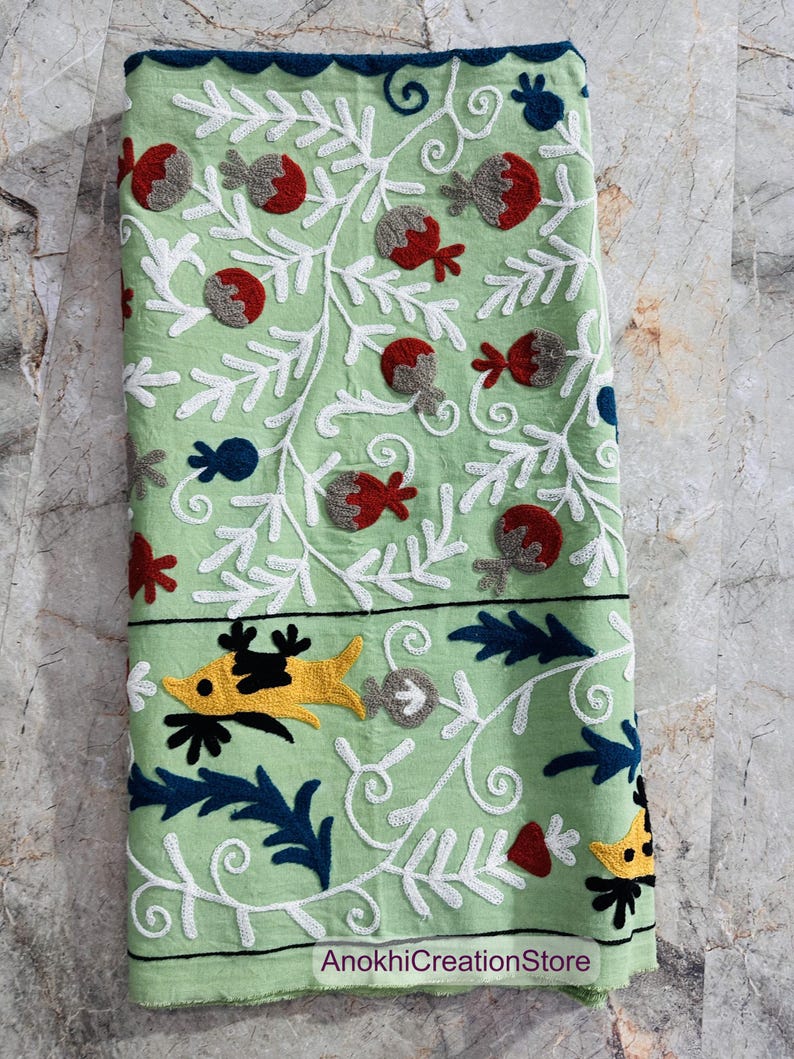 May include: A folded, light green textile with intricate embroidery. The design features white vines, red and gray floral elements, and yellow and black fish-like figures. The border is dark blue, and the text "AnokhiCreationStore" is visible.