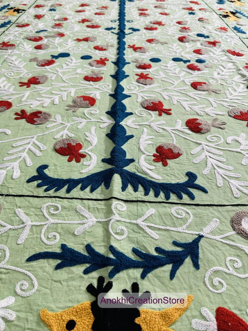 May include: A light green textile with intricate embroidery. The design features white, blue, red, and gray floral and leaf patterns. The text "AnokhiCreationStore" is visible in the lower right corner. The textile is likely a decorative item.
