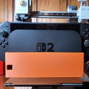 May include: A black Nintendo Switch console with a matching black stand and an orange base. The console has the text "D2" printed on it. The image is taken indoors with a wooden background.
