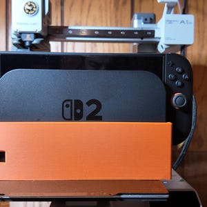 May include: A black Nintendo Switch console with a matching black stand and an orange base. The console has the text "D2" printed on it. The image is taken indoors with a wooden background.