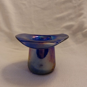May include: A small, iridescent blue glass vase shaped like a hat. The vase has a wide, flat brim and a tall, cylindrical base.