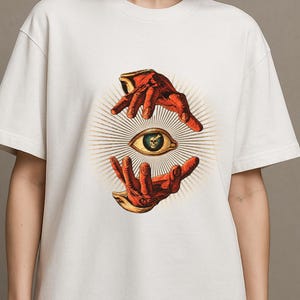 Op de afbeelding: Een off-white t-shirt met korte mouwen en een grafische print van twee rode handen die een centraal oog omvatten. In de pupil van het oog bevindt zich een kleine schedel. Het ontwerp is omringd door stralende gouden lijnen.
