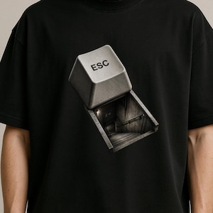 May include: A black short-sleeve t-shirt featuring a graphic print of a computer keyboard key labeled ESC. The key is partially lifted, revealing a dark, recessed staircase leading downward into a dimly lit, shadowed underground passage.