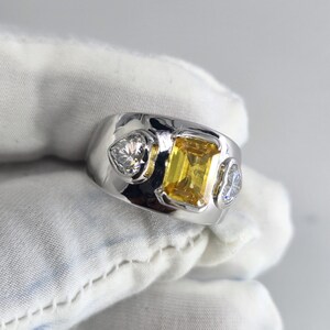 May include: A silver ring with a large, rectangular yellow gemstone in the center, flanked by two smaller, heart-shaped clear gemstones. The ring has a wide band and a polished finish.