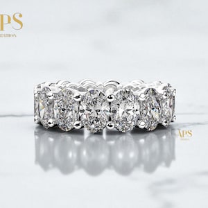 May include: A close-up of a diamond eternity ring. The ring features a series of oval-cut diamonds set in a white metal band. The diamonds sparkle, reflecting light. The ring is displayed on a reflective surface.