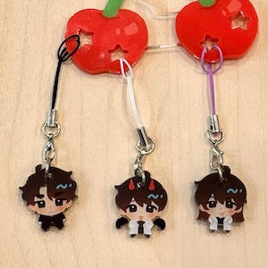 May include: A cherry-themed charm featuring two red cherries with star cutouts and green leaves. Each cherry has a small cartoon character charm attached. The characters have dark hair and wear black and white outfits. The charm is attached to a wooden surface.