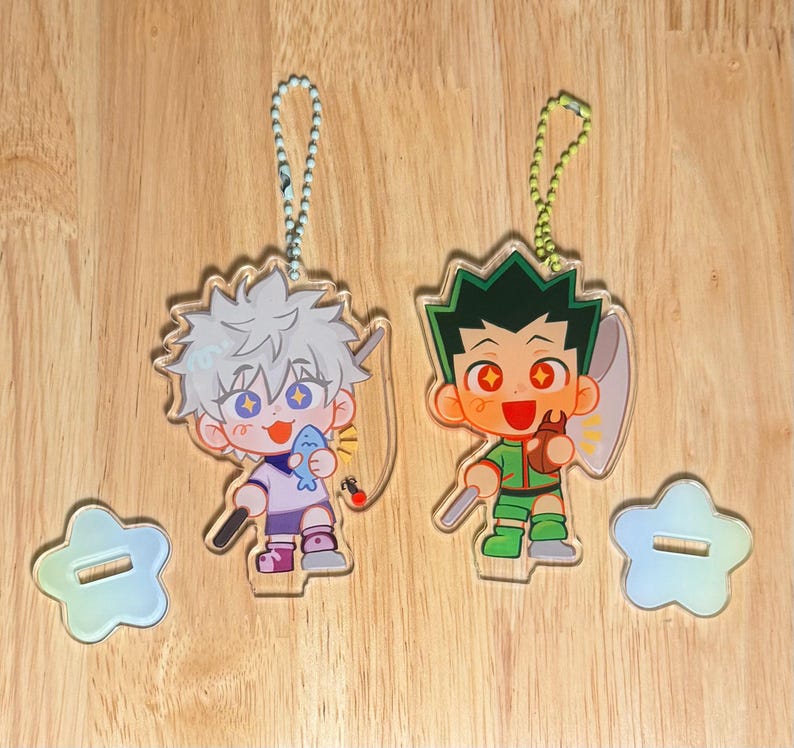 May include: Two acrylic anime character keychains. One keychain features a character with white hair holding a fish, the other features a character with green hair holding a fishing rod. Both have star-shaped stands.
