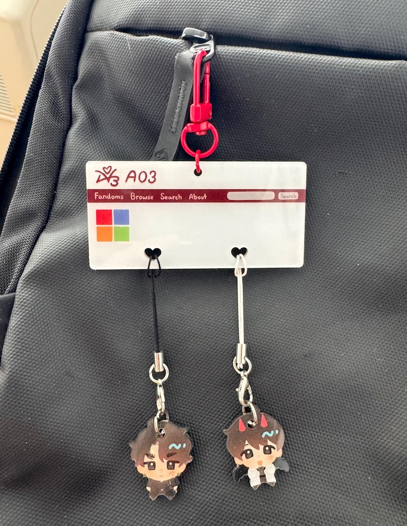 Puede incluir: Un charm de acr&iacute;lico blanco con el texto "A03" y "Fandoms Browse Search About" est&aacute; sujeto a una mochila negra con un mosquet&oacute;n rojo. Dos peque&ntilde;os amuletos de personajes cuelgan debajo. Los amuletos tienen un estilo de dibujos animados.