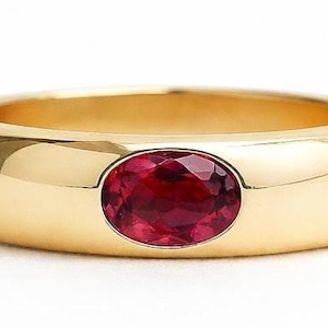 May include: A gold ring with a large, oval-shaped, red gemstone set in the center. The ring has a thick band and a polished finish, reflecting light. The gemstone is a vibrant, deep red color.