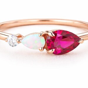 May include: Rose gold ring featuring three gemstones: a small, round, clear stone, a pear-shaped opal, and a large, pear-shaped ruby. The ring's band is thin and polished, with the gemstones set in a row.