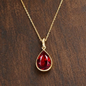 Op de afbeelding: Een gouden ketting met een druppelvormige rode edelsteen hanger. De hanger is gezet in een gepolijst gouden frame en hangt aan een fijne gouden ketting. De levendige rode steen heeft een gefacetteerd oppervlak dat licht reflecteert, wat zorgt voor een heldere, schitterende uitstraling in de gouden zetting.