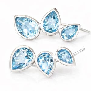 May include: Two pairs of silver earrings, each featuring three light blue, pear-shaped gemstones. The gemstones are set in a silver bezel, creating a cluster design. The earrings have a post-and-clutch closure.