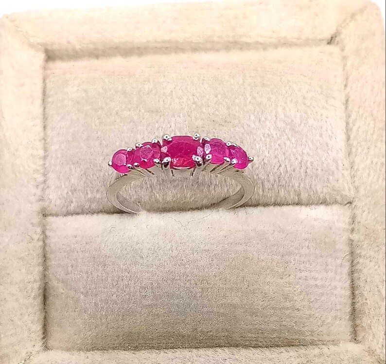 May include: A silver ring featuring a row of six vibrant, oval-cut rubies. The rubies are a deep, rich pink color, set closely together. The ring is displayed on a soft, off-white surface.