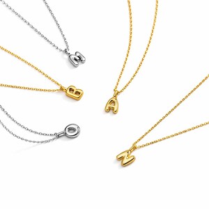 May include: A collection of gold and silver-toned initial necklaces. The necklaces feature the letters A, B, M, O, and N. Each letter is a pendant on a delicate chain. The pendants have a rounded, bubble-like design.